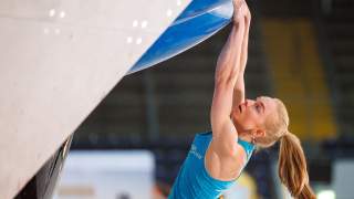 Frau mit Pferdeschwanz klettert an einer Indoor-Kletterwand und greift nach einem blauen Griff. Sie trägt ein blaues ärmelloses Sportshirt.