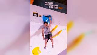Ein Boulderer hängt an einem blauen Griff an einer Kletterwand bei der Deutschen Meisterschaft Bouldern 2025. Die Wand ist mit weiteren bunten Griffen in Gelb und Beschriftungen versehen.