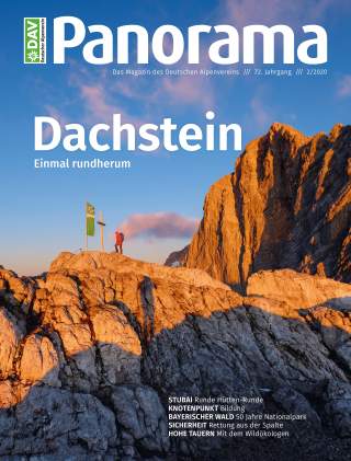 Ein Wanderer steht auf einem Felsen am Gipfel bei Sonnenuntergang neben einer Fahne und einem Gipfelkreuz, umgeben von felsigen Bergen unter blauem Himmel.