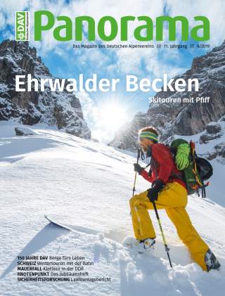 Ein Skitourengeher in gelber Hose und roter Jacke steigt mit Skistöcken in tiefem Schnee zwischen felsigen, schneebedeckten Bergen bei Sonnenschein auf.