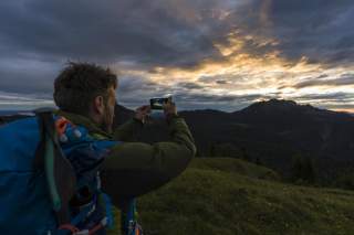 Ein Wanderer mit Rucksack fotografiert bei Sonnenuntergang eine Berglandschaft mit bewölktem Himmel.
